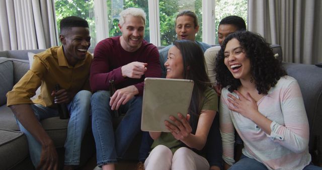 A diverse group of friends are sitting on a couch at home, laughing and using a tablet together. This image is ideal for advertising social media apps, friendship-focused campaigns, mobile technology products, and lifestyle blogs about home entertainment and social gatherings.
