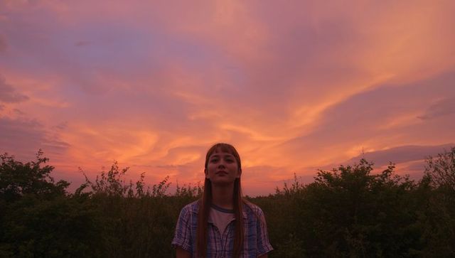 Young woman standing and gazing at dramatic pink-orange sunset over meadow silhouette