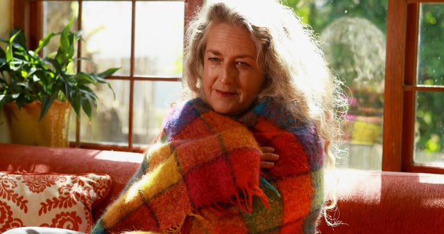 Senior woman wrapped in colorful plaid blanket, basking in natural light from large windows. Relaxing on a red sofa with decorative cushions, surrounded by a warm and inviting home atmosphere. Ideal for uses in home decor, lifestyle, senior living, and comfort-related projects.