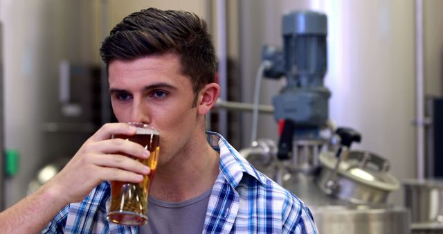 This image depicts a man in a casual plaid shirt sipping a glass of beer in a brewery. Brewing equipment is visible in the background. It can be used for marketing beer brands, brewery tours, or articles on brewing and beer tasting.