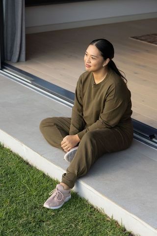 Mid-adult asian woman sitting on doorstep in olive loungewear and pink sneakers, relaxing