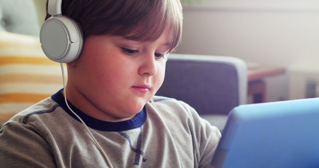 Young boy using headphones while engaged with a tablet at home. Perfect for educational articles, technology products marketed to children, articles on screen time for kids, or feature pieces on modern child entertainment strategies. Can illustrate homeschooling or digital learning during leisure time.