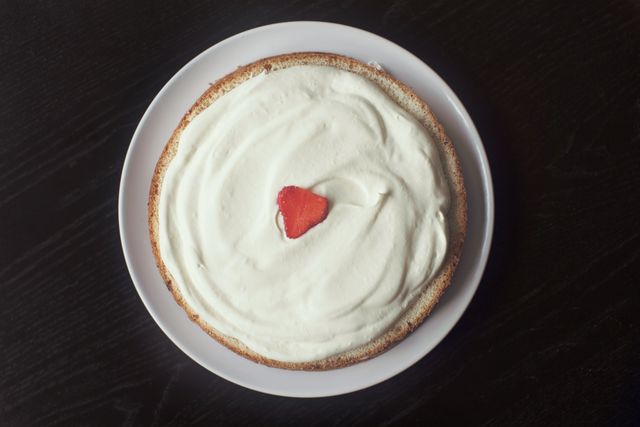 Round cake topped with smooth whipped cream and a single strawberry slice in the center, displayed on a white plate against dark background. Ideal for use in food blogs, recipe websites, and dessert-themed marketing materials.