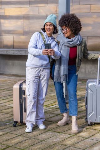Diverse female friends checking smartphone while traveling with rolling suitcases