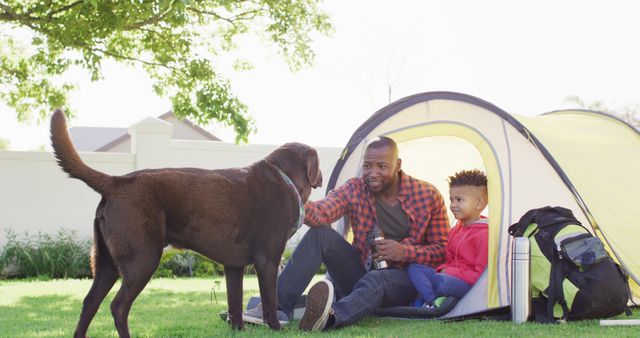 Father and son sitting at tent's entrance on lawn with dog nearby. Father in plaid shirt bonding with son. Appears to be back lawn of a house with green shrubs and tree overhead. Ideal for family-oriented brands, outdoor camping promotions, pet care products, and lifestyle advertisements.