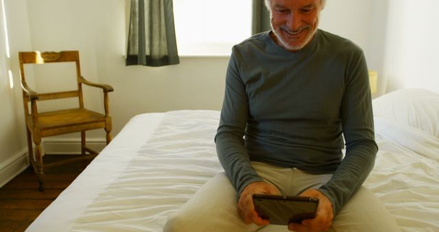 Elderly man sits on bed in bright, airy bedroom while using a tablet. The man is smiling and looks relaxed, symbolizing a positive interaction with modern technology. Ideal for concepts related to seniors and technology, digital literacy for the elderly, and casual home settings.