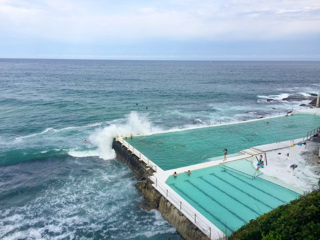 This image shows a large outdoor swimming pool situated right next to the ocean. Ocean waves are crashing against the edge of the pool with people swimming and relaxing nearby. Perfect for use in travel brochures, vacation advertisements, resort promotions, and articles focusing on coastal and seaside locations.