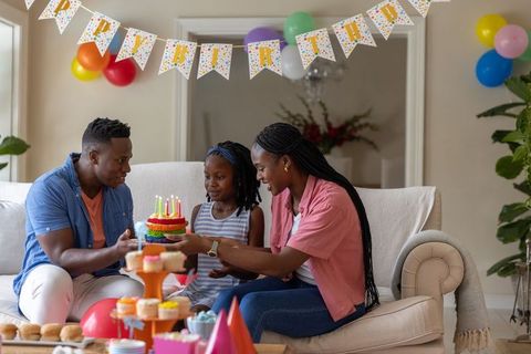 Family celebrating daughter birthday at home with rainbow cake, balloons, candles