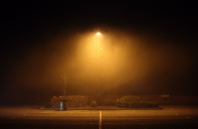 An empty parking lot at night illuminated by a single streetlight with thick fog surrounding the area. This image evokes feelings of solitude and eeriness, making it suitable for themes of mystery, suspense, urban exploration, and nighttime scenery. Perfect for website headers, blog posts related to urban landscapes, mysterious storytelling, or illustrating concepts of isolation and serenity.