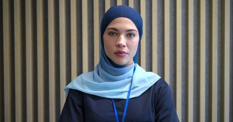 Confident businesswoman wearing light blue hijab and navy top with lanyard in office with woodslats