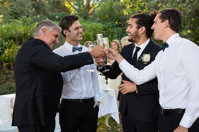 Groom and guests toasting with champagne glasses in an outdoor park setting. Ideal for use in wedding invitations, celebration announcements, event planning websites, and articles about wedding traditions and celebrations.