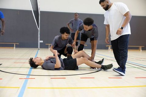 Teenage volleyball player lying injured on gym court while teammates and coach giving aid