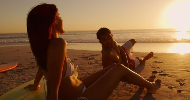 Biracial couple enjoys a sunset on the beach, with copy space. They share a relaxing moment after surfing, highlighting a romantic outdoor setting.