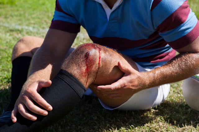 Rugby player sitting on grass field with a bleeding knee injury. Ideal for use in articles about sports injuries, healthcare, first aid, and athletic safety. Can be used in educational materials, injury prevention campaigns, and sports-related content.