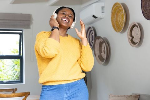 Young african american woman enjoying music at home in yellow sweater with wireless headphones
