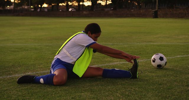 Soccer player stretching legs on a grassy field during sunset, preparing for practice or a match. Ideal for use in sports training materials, fitness blogs, healthy lifestyle promotions, and athletic apparel advertisements.