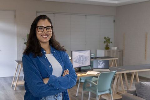 Confident woman architect standing in modern design studio with cad screens and laptop