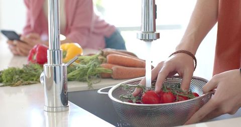 Teen washing vine tomatoes under kitchen faucet while family prepping fresh vegetables