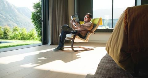 Asian man relaxing in armchair reading book by glass doors