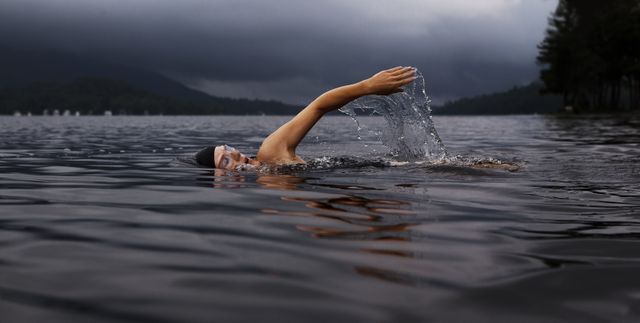 The photo captures a man swimming in a calm lake under an overcast sky with dark storm clouds gathering in the distance. The water reflects the approaching weather creating a dramatic and intense atmosphere. This image is suitable for use in outdoor fitness promotions, adventure blogs, environmental campaigns, and inspiring stories about perseverance and challenging weather conditions.