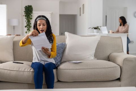 Teenage daughter opening exciting letter on sofa while mother working on laptop in background