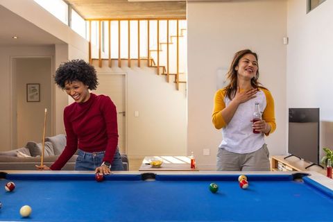 Diverse women laughing and playing pool in modern living room with drinks