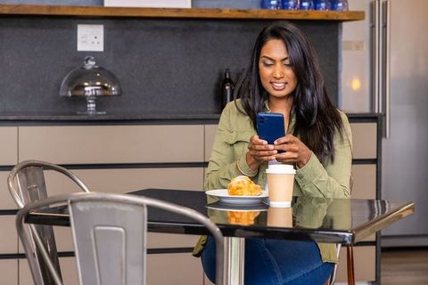 Indian woman using smartphone while enjoying coffee and croissant in modern casual kitchen
