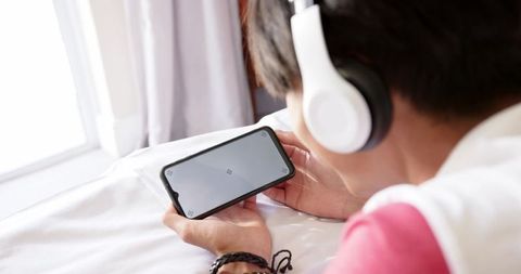 Teenage asian boy lying on bed watching videos on smartphone while wearing headphones