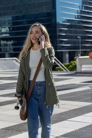 Young woman walking and talking on phone carrying skateboard in sunlit urban plaza