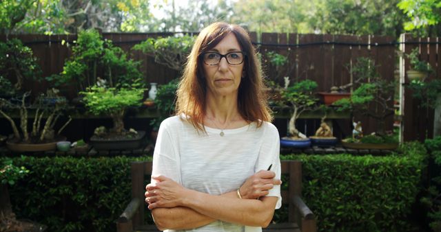 A middle-aged woman with glasses stands with crossed arms in her garden. She is wearing casual clothes and has a composed, serious expression. The background features a variety of garden plants and a wooden fence. This image is ideal for use in topics related to gardening, lifestyle, aging, confidence, and outdoor activities.