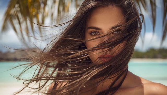 Gazing brunette with windblown hair at turquoise lagoon beach, sunlit tropical closeup