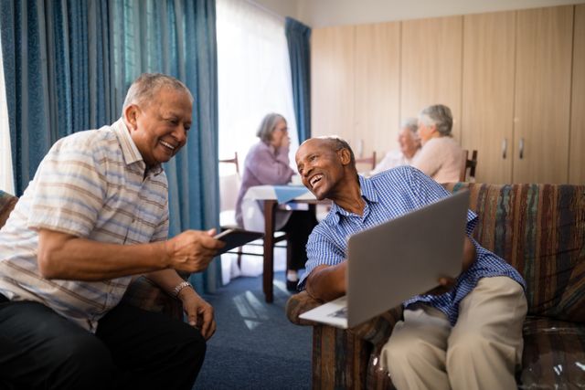 Senior men sitting on a sofa in a nursing home, one showing a laptop to his friend. They are smiling and enjoying each other's company. In the background, other elderly people are sitting and conversing. This image can be used for promoting elderly care facilities, showcasing the positive aspects of retirement living, or highlighting the use of technology among seniors.