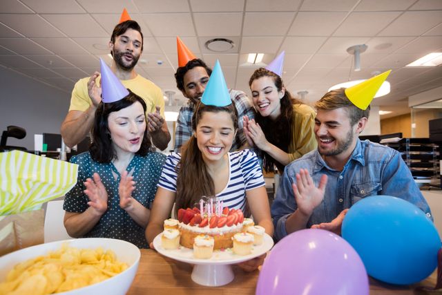 Group of colleagues celebrating a birthday in an office environment. They are gathered around a table with a birthday cake, cupcakes, and balloons, wearing party hats and smiling. This image can be used for promoting workplace culture, team building, and corporate events.