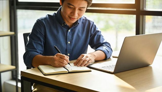 Asian man writing in notebook at sunlit minimalist workspace with laptop and natural light