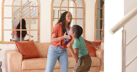 African american mom and teen daughter laughing and hugging in cozy living room by sofa