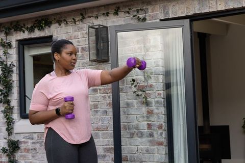 African american woman lifting dumbbells on backyard patio wearing pink shirt and smartwatch