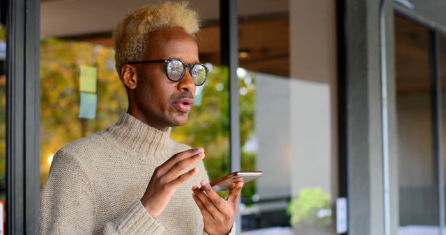 Man using voice command feature on smartphone. Wearing glasses and beige sweater. Standing outside building, framed by windows and plants in background. Perfect for illustrating technology, modern communication, app usage, and lifestyle concepts.