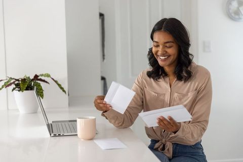 Young woman smiling and sorting mail at modern kitchen counter with laptop and mug