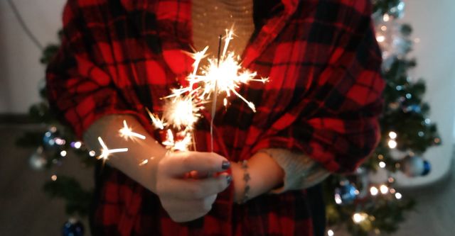 Image shows person holding lit sparklers with a Christmas tree and ornaments in the background. Ideal for use in holiday-themed content, festive event promotions, seasonal greeting cards, and decorating inspirations.