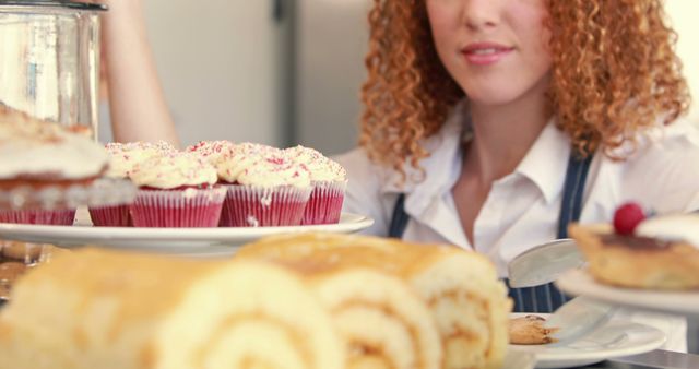 This image shows a woman with curly hair standing behind a counter with various pastries and cupcakes on display. Perfect for illustrating bakery shop promotions, dessert recipes, or lifestyle blogs focusing on culinary delights.