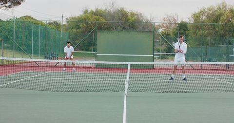 Men playing tennis on outdoor hard court holding rackets wearing white kit returning serve