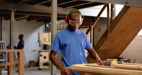 Carpenter sanding wooden planks wearing safety goggles and earmuffs at busy woodworking shop