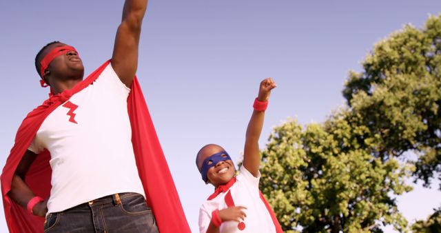 Two African American individuals, a parent and child, are dressed in superhero costumes with capes and masks, striking triumphant poses outdoors, with copy space. Their playful expressions and superhero attire suggest a fun, imaginative day spent together.