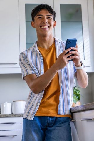 Smiling asian man holding blue smartphone in modern kitchen, casual smartwatch lifestyle