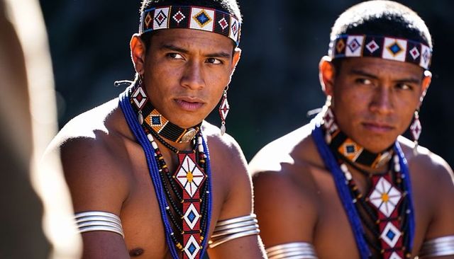 Indigenous men wearing beaded regalia and metal armbands during outdoor ceremony