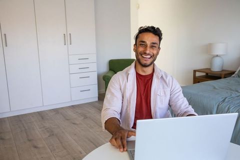 Smiling South Asian Man Using Laptop in Cozy Modern Bedroom