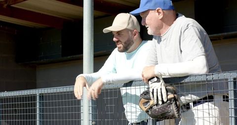 Veteran and younger teammate leaning on dugout rail holding baseball glove wearing caps