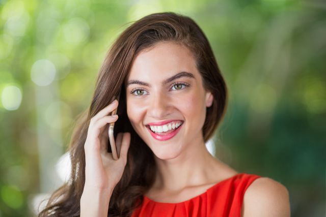 This image shows a young woman smiling while talking on her mobile phone outdoors. The background is blurred with greenery, suggesting a natural setting. This image can be used for promoting mobile communication services, lifestyle blogs, technology advertisements, or any content related to happiness and casual conversations.