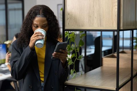 African-american professional sipping coffee and checking smartphone in coworking office