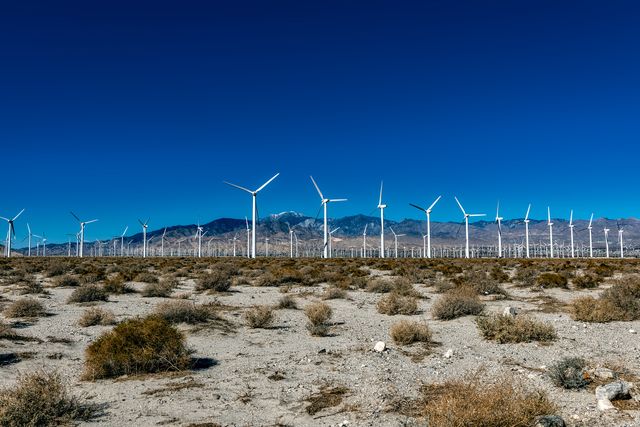 Wind turbines are prominently standing in a desert landscape with a clear blue sky and mountains in the background. Ideal for use in articles, publications, or websites focusing on renewable energy, sustainability, environmental conservation, or desert landscapes. The clear sky and open desert emphasize the power and potential of renewable energy sources.